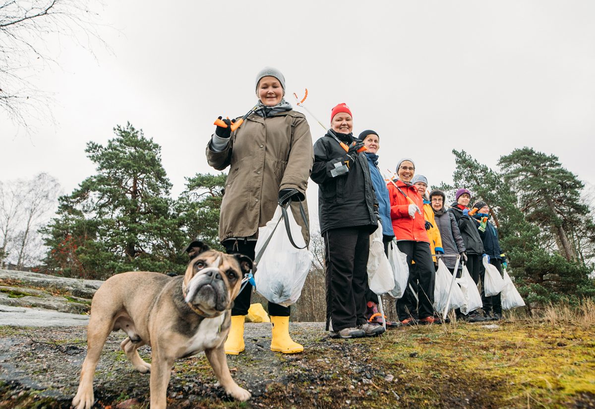 Vapaaehtoiset siivoavat rantaa roskista. He seisovat rivissä roskapussien kanssa ja heillä on koira mukanaan.